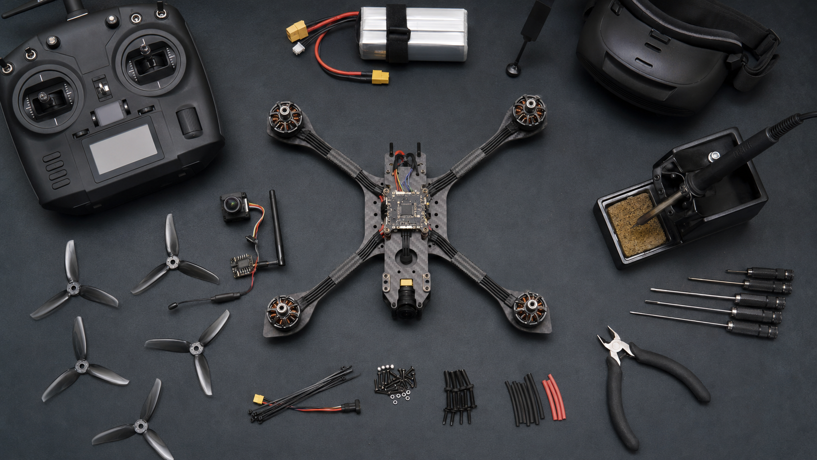 FPV drone build kit parts arranged on a workbench, including a carbon frame, motors, controller, goggles, battery and soldering tools.