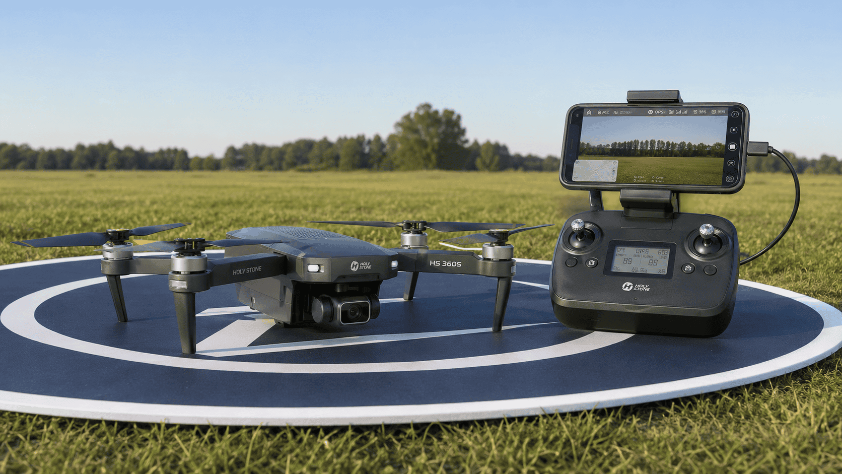 Holy Stone HS360S beside its controller and a phone displaying the HS FLY app at an outdoor launch site
