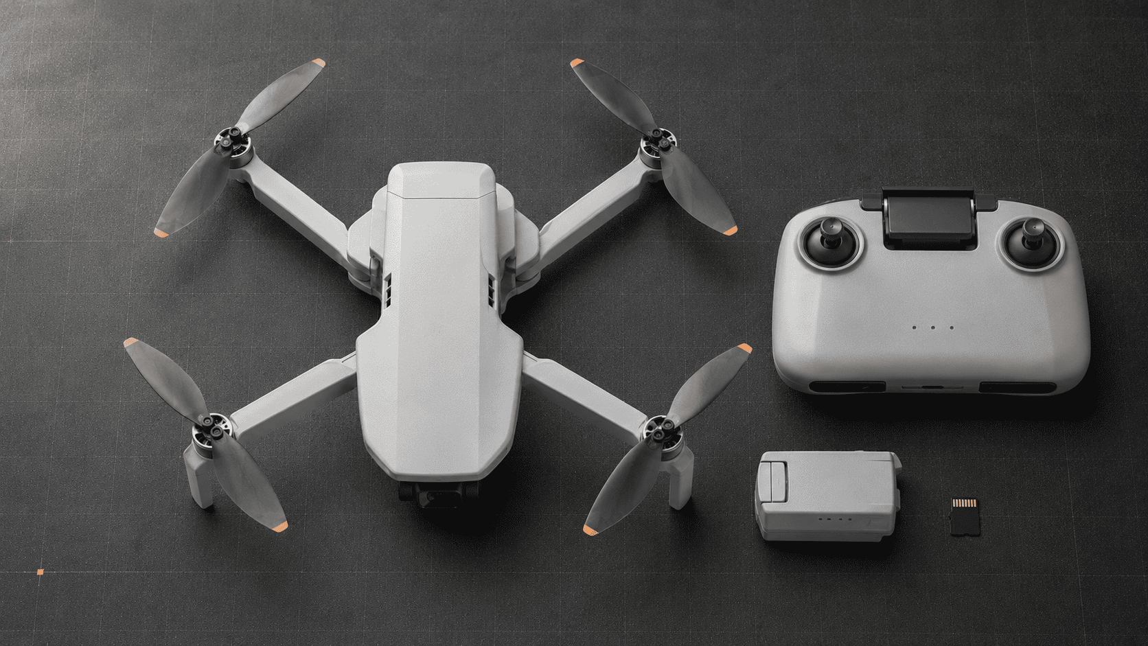 Compact gray folding drone beside its controller, battery, and microSD card on a technical work surface.