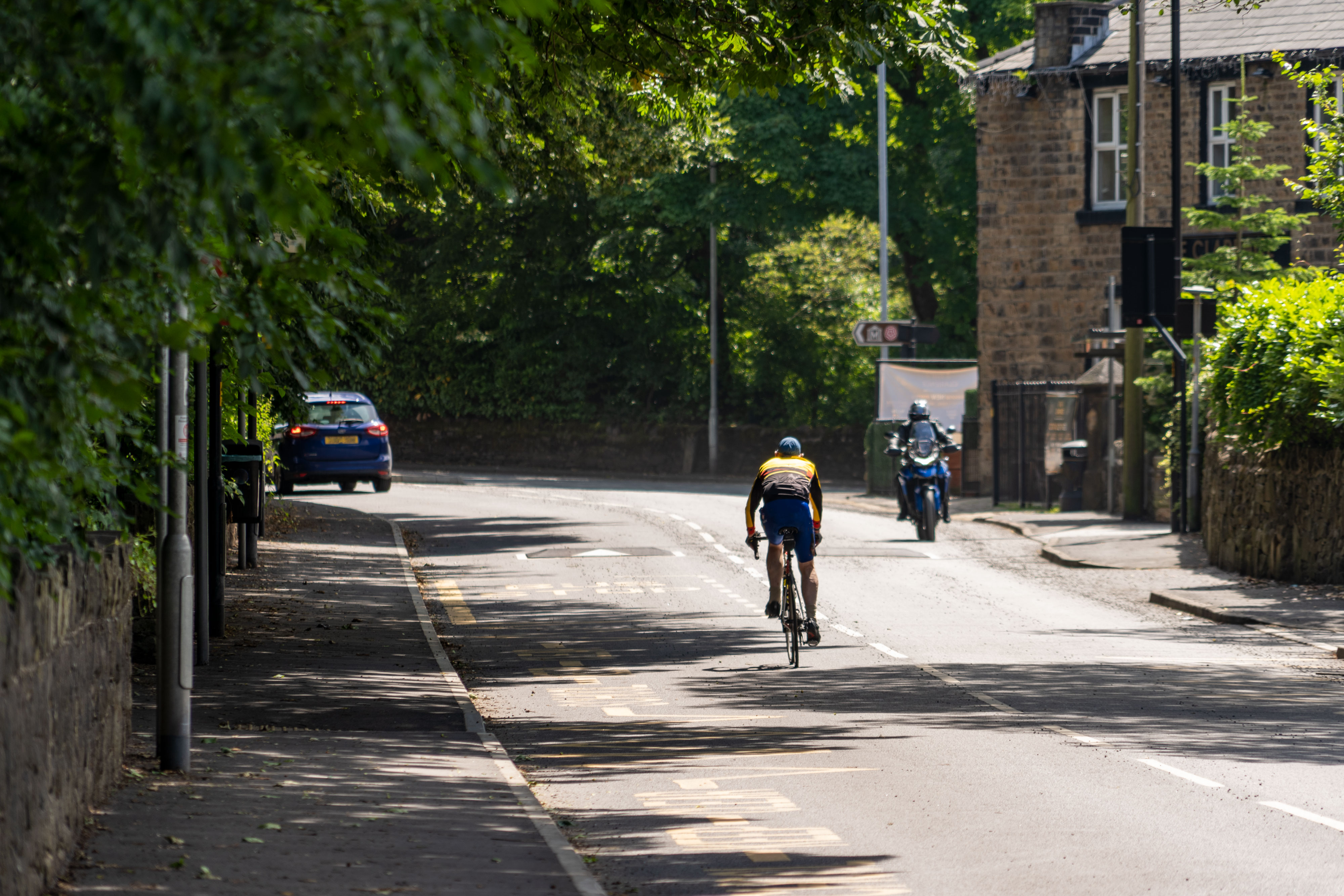 A cyclist approaching a bend with traffic, roadside walls, buildings and overhanging trees.