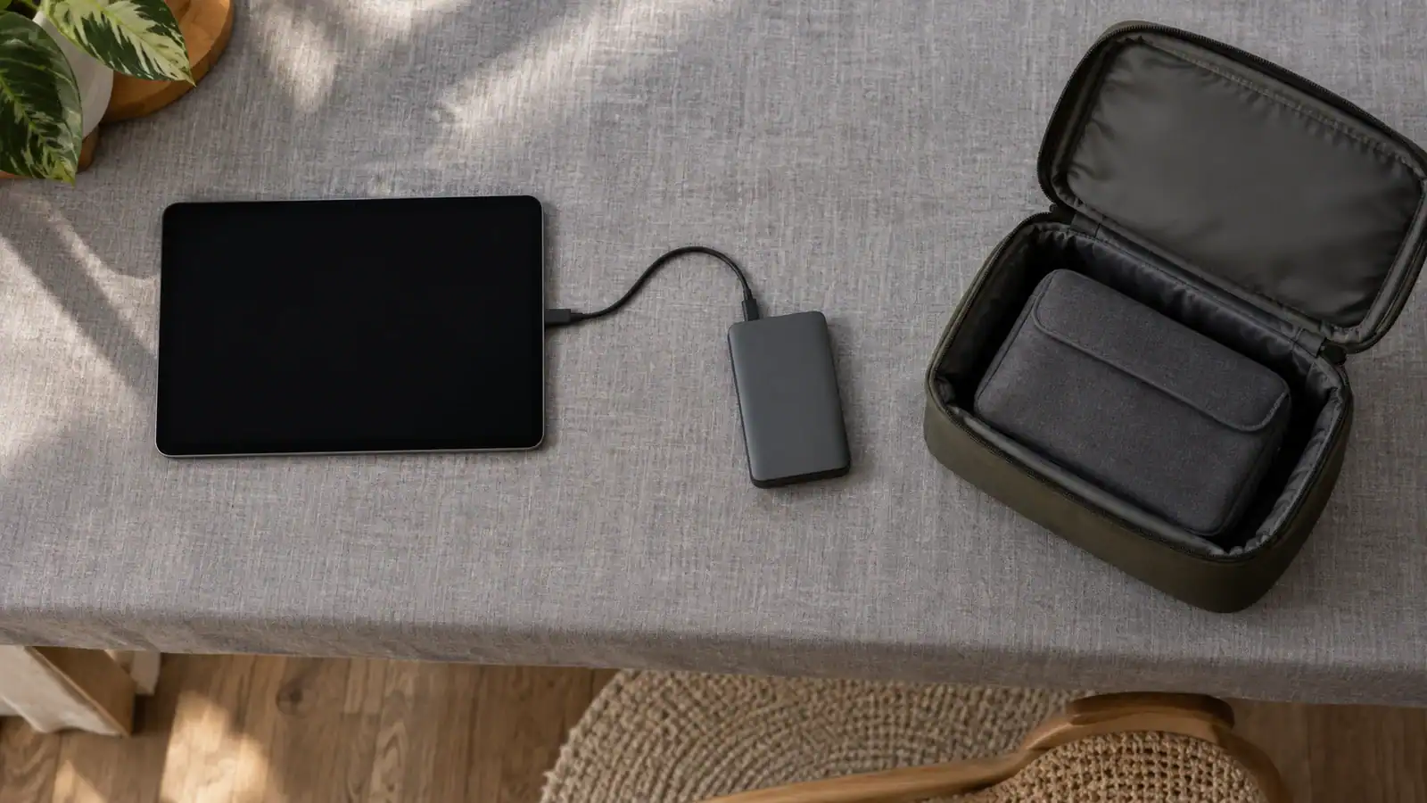 Overhead view of a dark-screen tablet connected to a portable drive beside an open padded equipment pouch on a gray fabric-covered table.