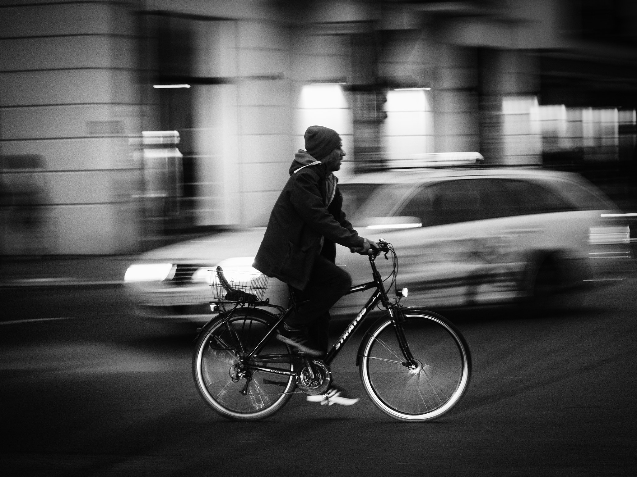 Black-and-white photograph of a cyclist passing a car and buildings blurred by camera movement.
