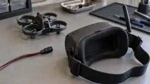 Unbranded FPV goggles with a dark screen and disconnected power lead on a gray workbench, with a small ducted quadcopter softly blurred behind.