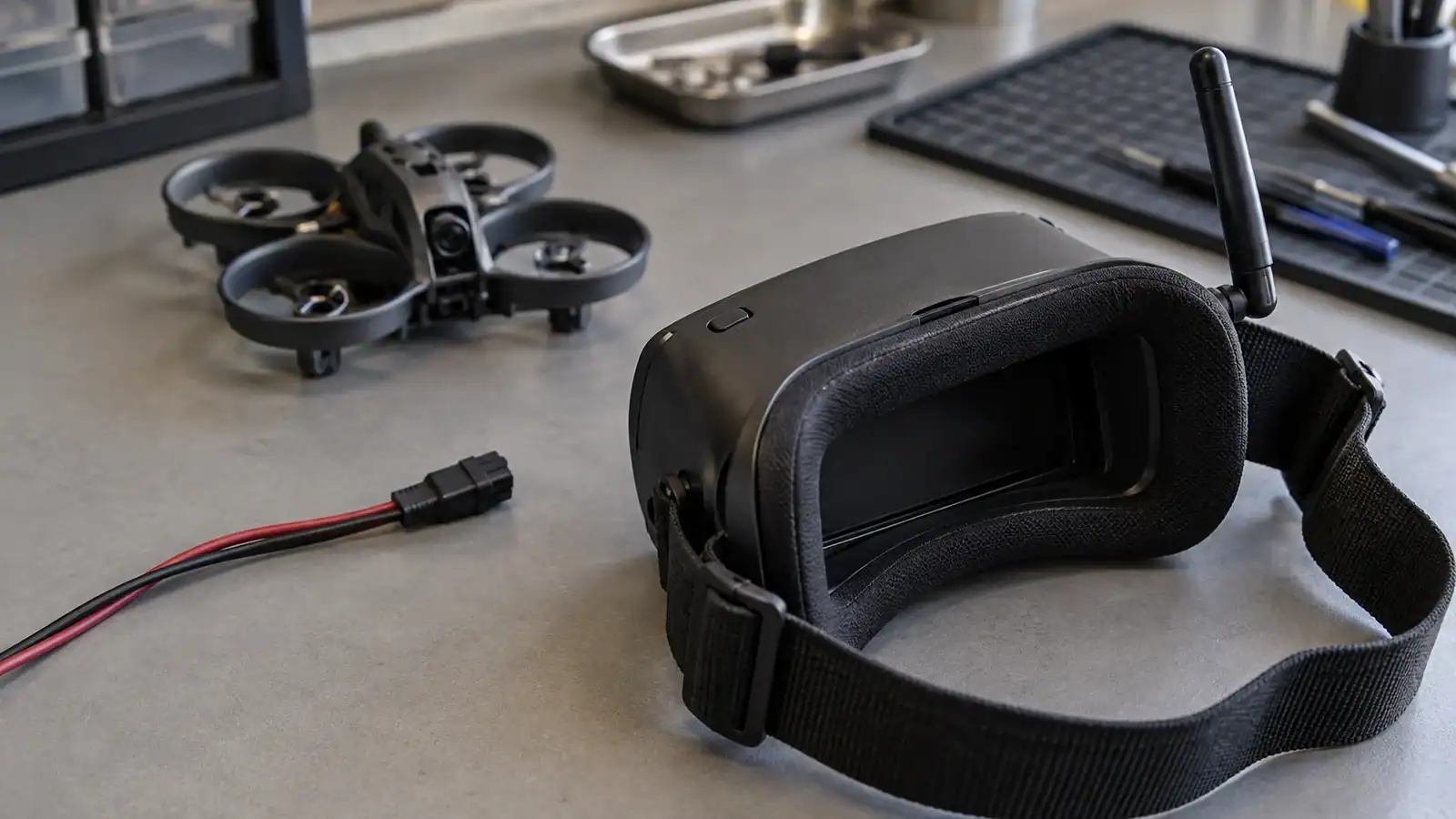 Unbranded FPV goggles with a dark screen and disconnected power lead on a gray workbench, with a small ducted quadcopter softly blurred behind.