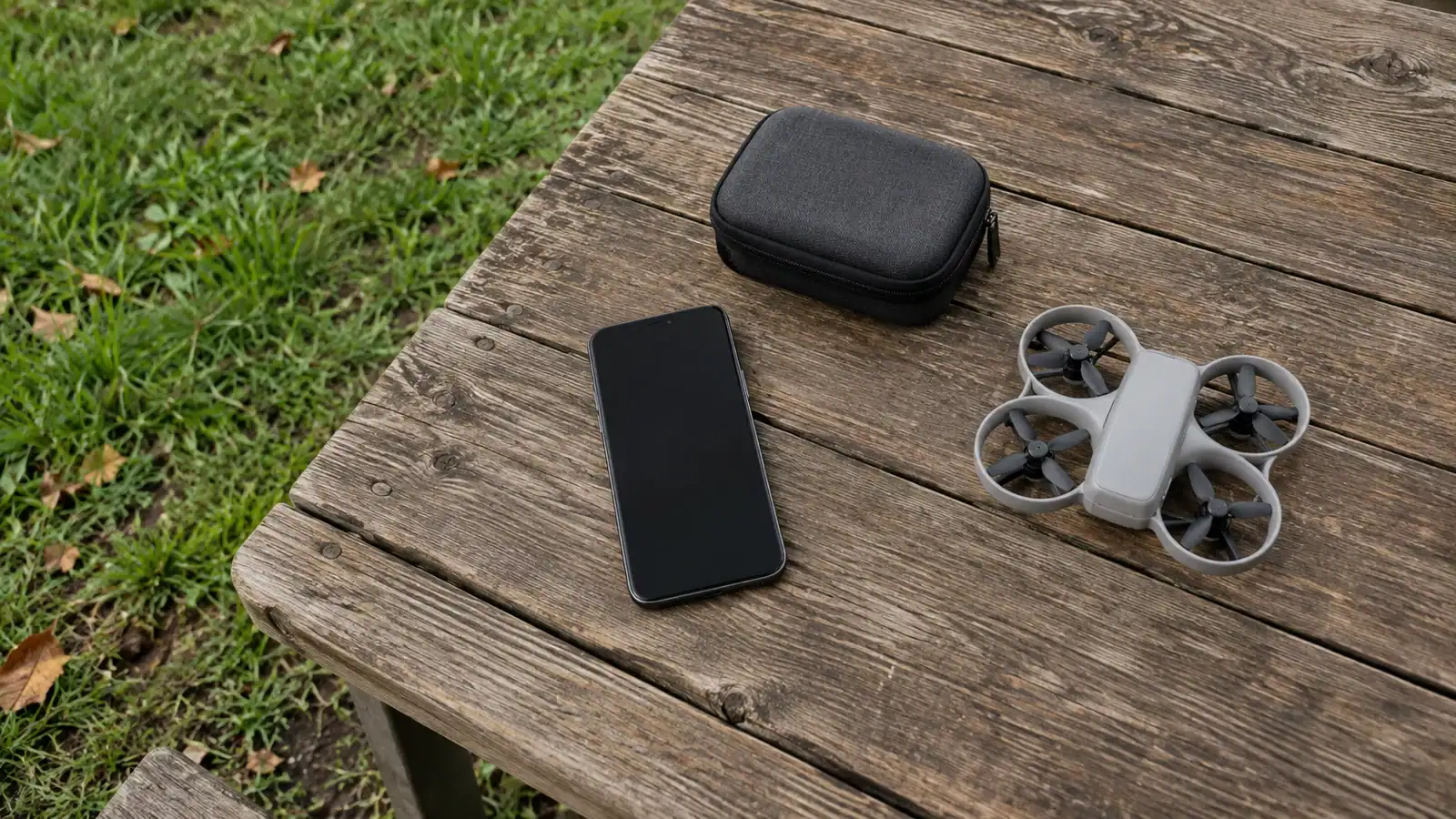 A dark-screen smartphone, plain carrying case, and compact unbranded drone sit separately on a wooden outdoor table beside grass.